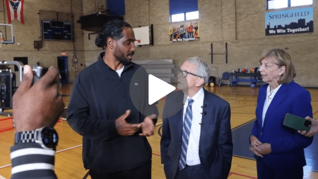 Three adults conversing in a gymnasium, with two holding recording devices.