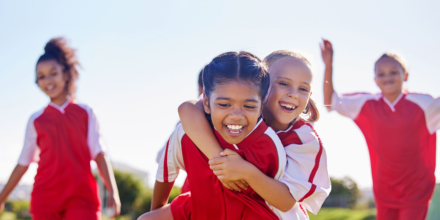 Two smiling children in soccer jerseys embracing on a field.