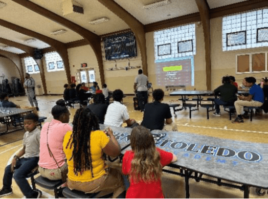 Adults and children watch a presentation in a gymnasium.