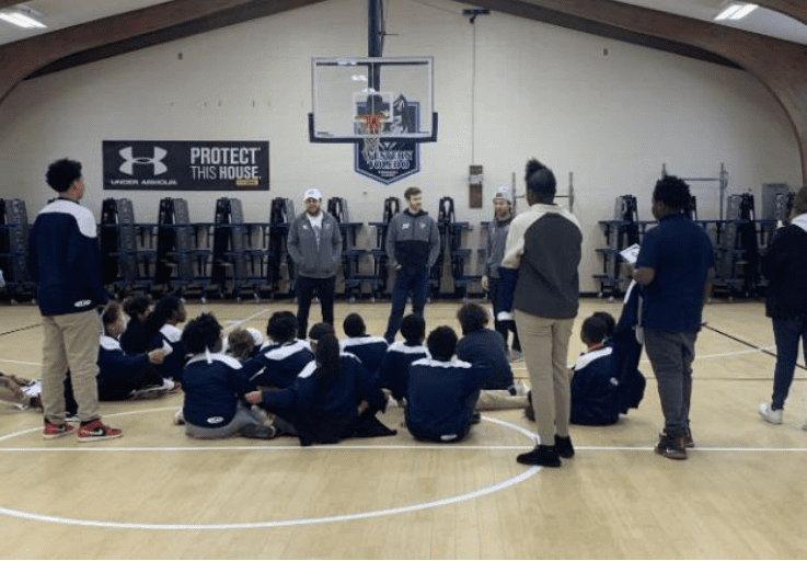 Adults addressing children sitting on a gymnasium floor.