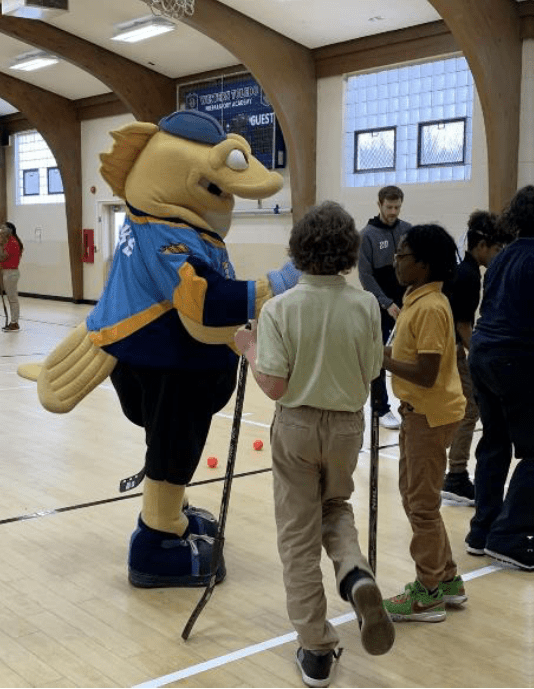 Mascot, dressed as a fish, interacts with children holding hockey sticks.