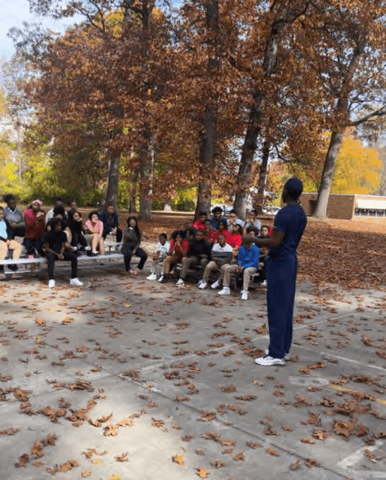 Adult speaking to children seated on bleachers outdoors.