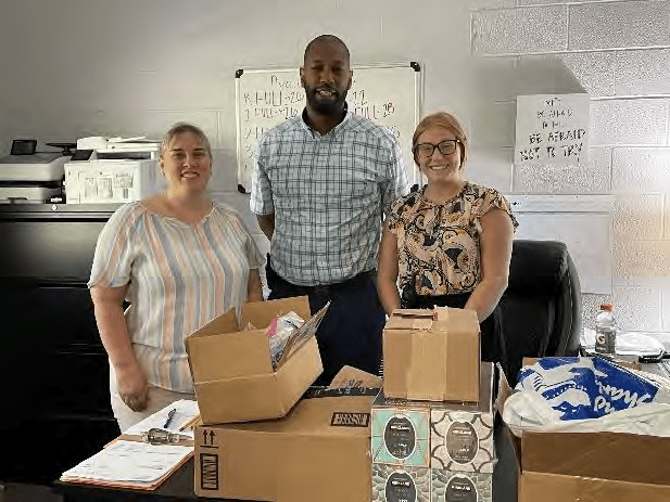Three adults stand smiling behind a table with boxes.