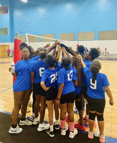Children in blue shirts huddle on a volleyball court.